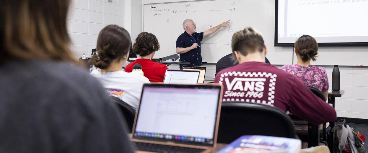 A professor stands in front of a classroom of students, pointing at a whiteboard as he teaches. We see everything from the back of the classroom so we get a view of some students backs and an open laptop as a student takes notes. 