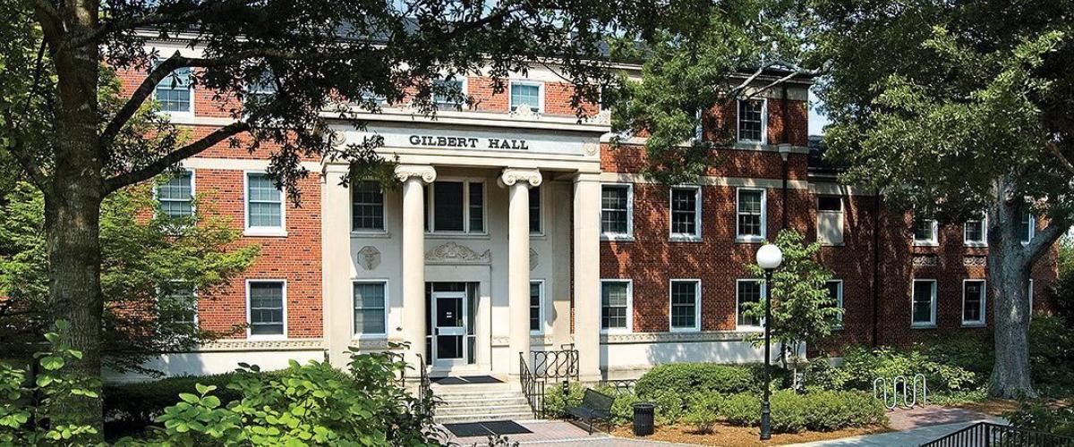 The front of Gilbert Hall, a brick building with white columns at the front and many windows with white trim. The building is surrounded with greenery. 