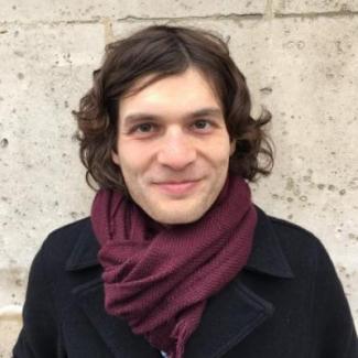 Portrait of Dr. Jon Forrest in a black coat and burgundy scarf standing in front of brick wall