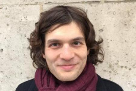 Portrait of Dr. Jon Forrest in a black coat and burgundy scarf standing in front of brick wall